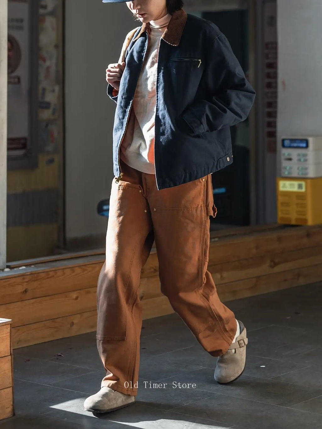 A person wearing a navy blue jacket with brown collar details, brown wide-leg cargo pants, a white turtleneck, and white slip-on shoes, walking in an indoor setting.