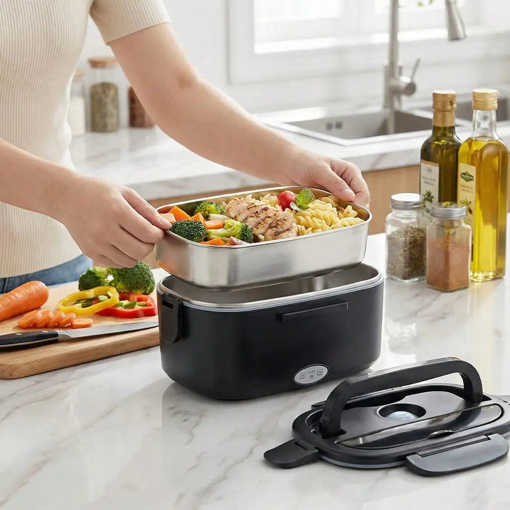 A black electric heated lunch box with a stainless steel tray filled with pasta, chicken, broccoli, and carrots, shown on a kitchen counter with a person lifting the tray. Cooking oil bottles, spices, and a cutting board with vegetables are visible in the background.