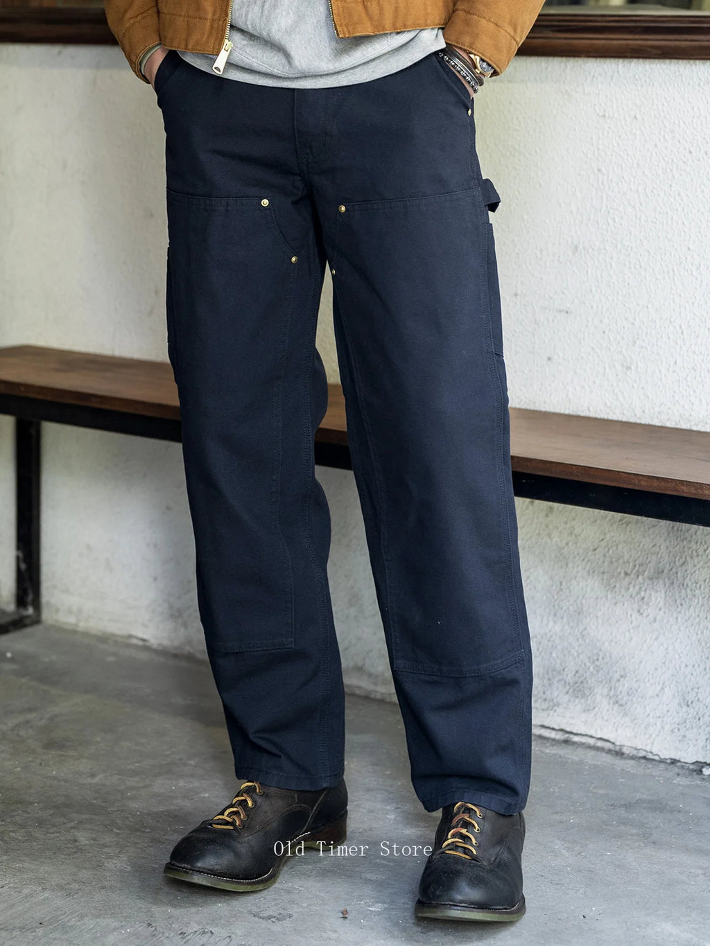 A person wearing dark navy work pants with multiple utility pockets, styled with a tan jacket, gray shirt, and dark brown boots, standing in front of a white wall and wooden bench