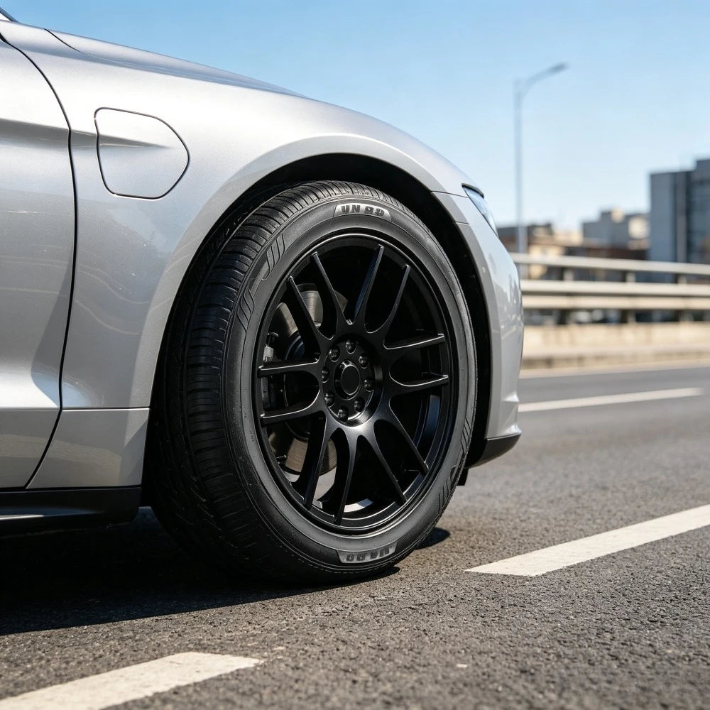 A close-up shot of a silver car's front wheel featuring a black multi-spoke alloy wheel and a black Pirelli tire, set on an asphalt road with a blurred city background.