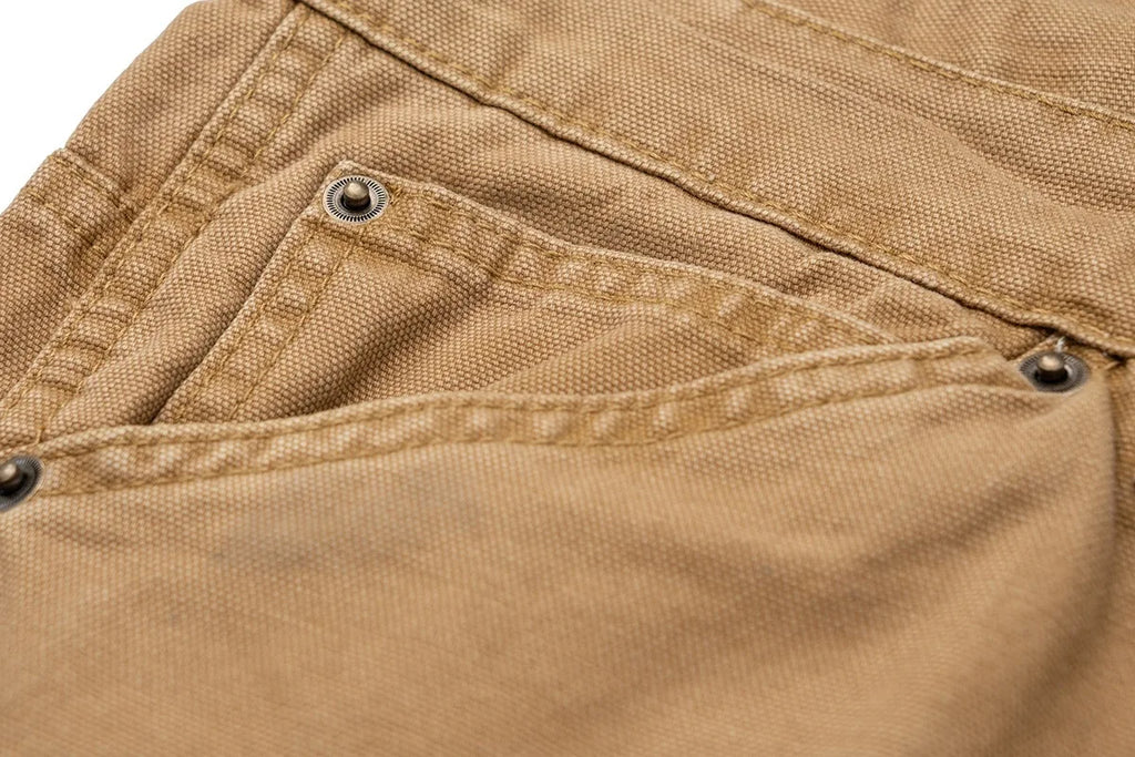 Close-up of a tan canvas garment, likely a jacket or overshirt, showing a pocket with metal rivets and stitched seams.