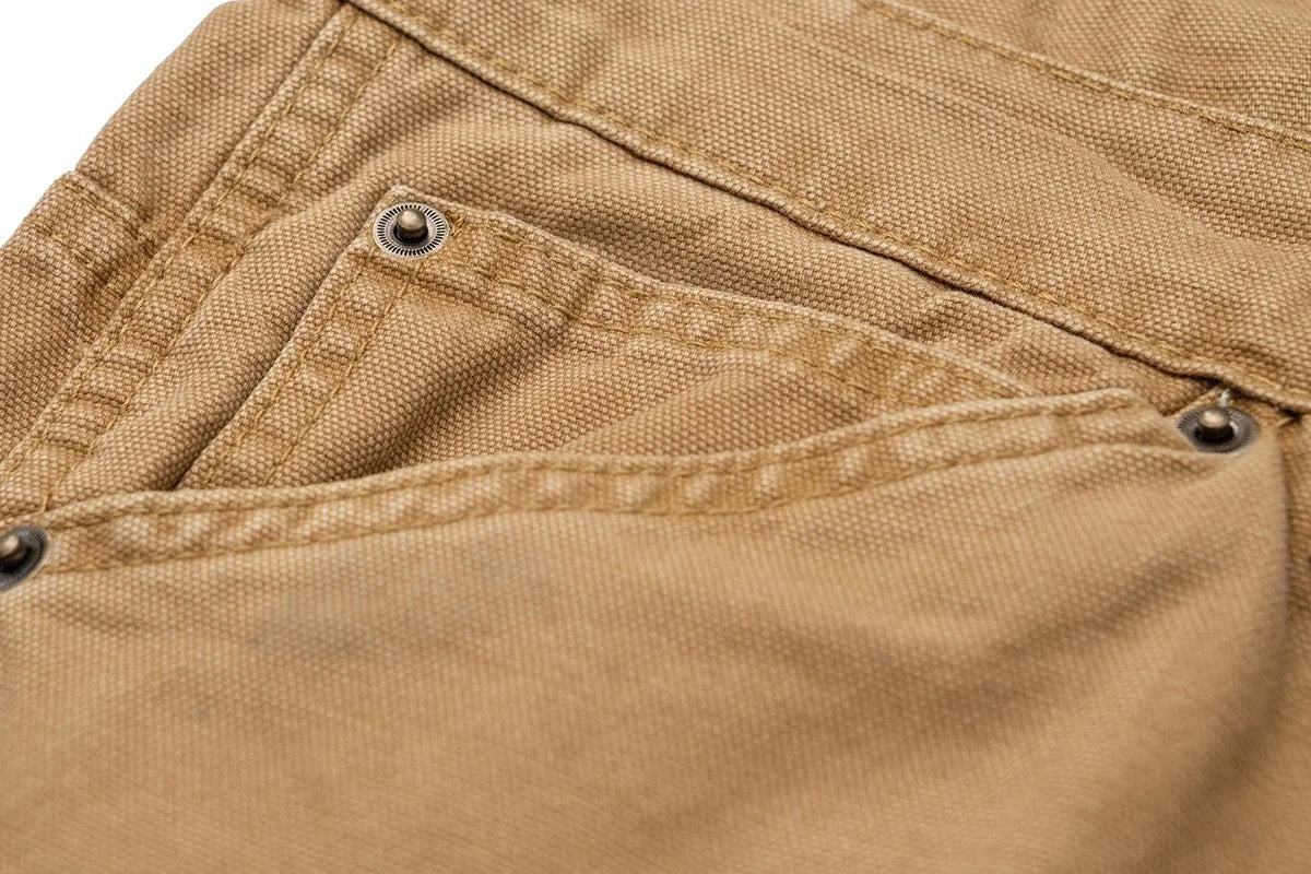 Close-up of a tan canvas garment, likely a jacket or overshirt, showing a pocket with metal rivets and stitched seams.