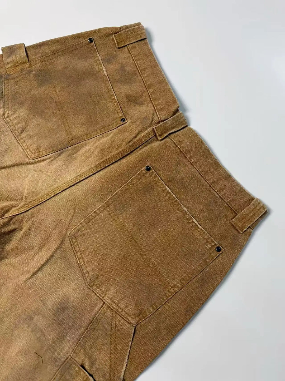 A close-up, diagonal view of a pair of brown canvas work pants showing the waistband, belt loops, and large front pockets against a plain light background.