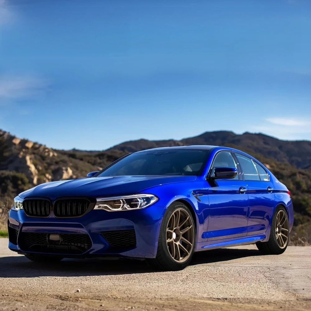 A blue BMW M5 sedan photographed outdoors on a dirt road with hills and a blue sky in the background.