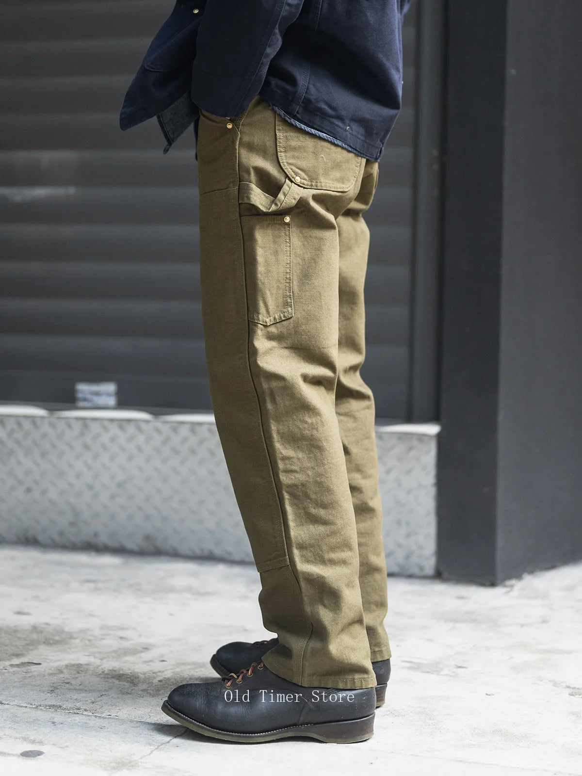 A person shown from the waist down wearing olive green canvas cargo pants, a dark navy shirt, and black leather shoes, standing on a concrete surface in front of a dark metal shutter.