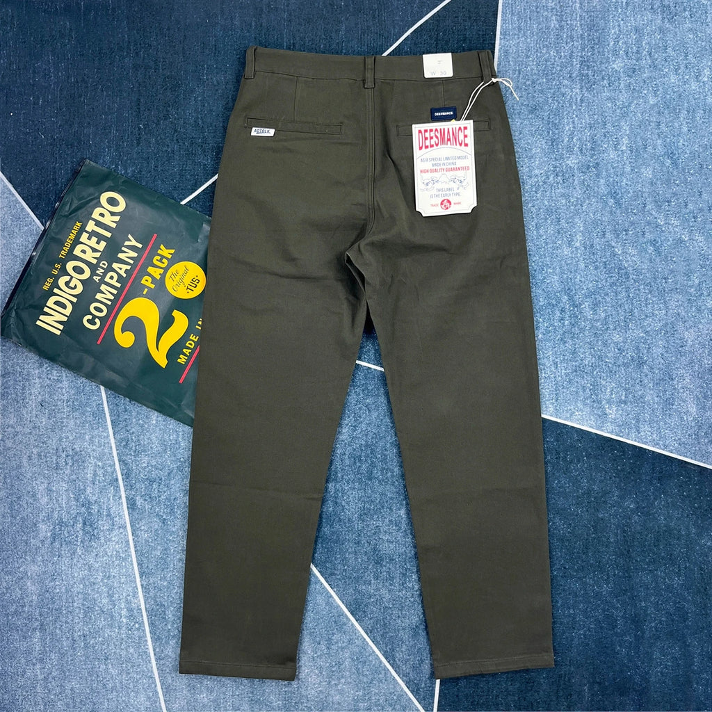 A pair of dark olive green chino pants shown from the back, laid flat on a blue patterned carpet next to a green and yellow Indigo Retro Company 2-pack tag.
