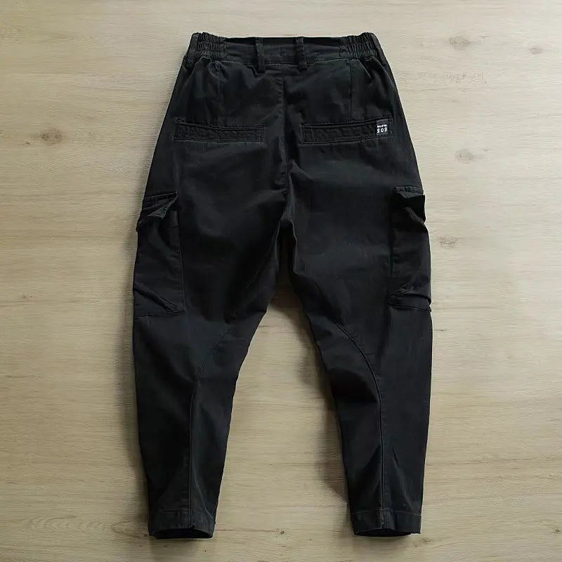 Black cargo pants with an elastic waistband, laid flat on a light wood floor. The back view shows cargo pockets and a small white brand tag on the right pocket.