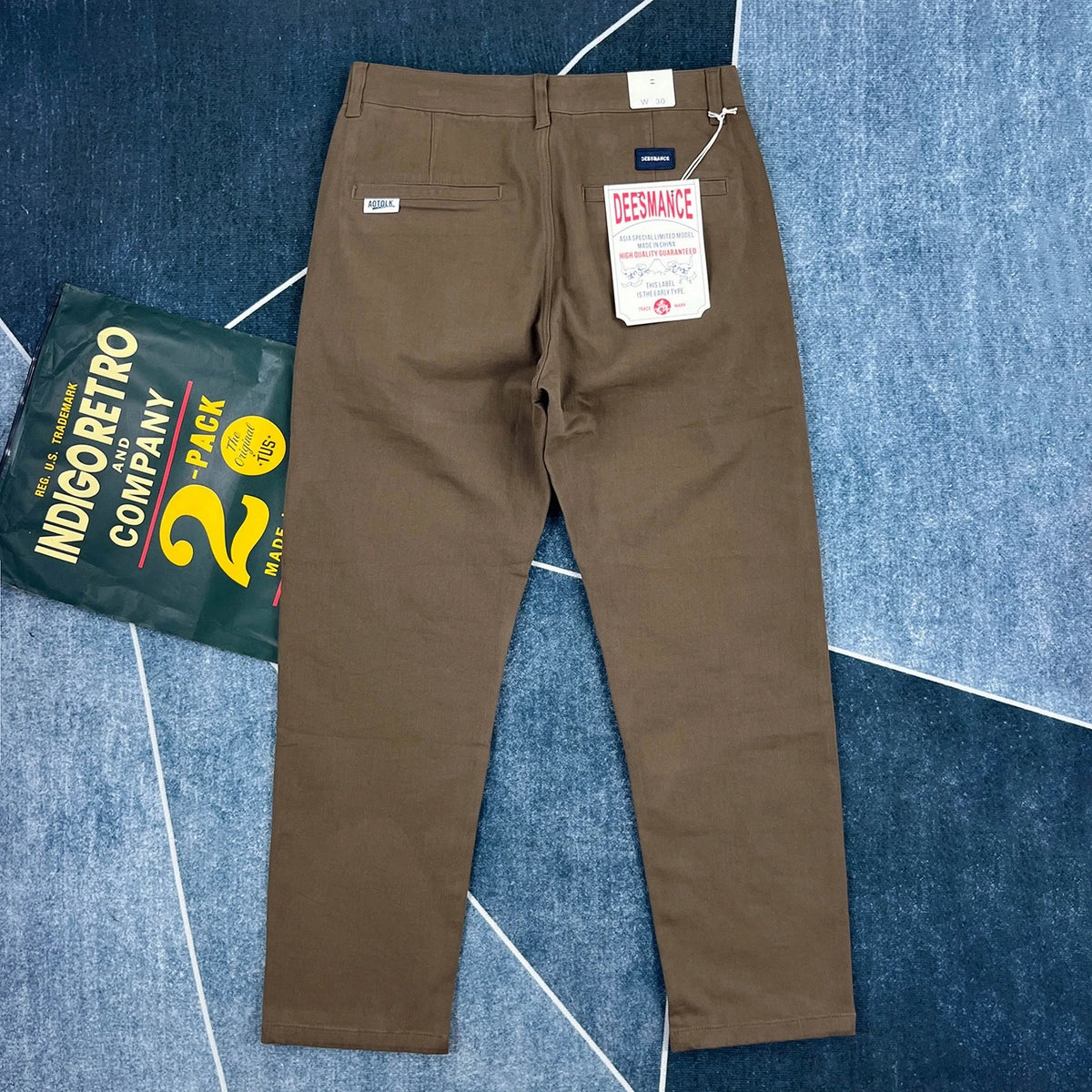 A pair of brown chino pants shown flat on a blue patterned carpet, with the back pockets, waistband, and hangtag visible. A green 'Indigo Retro Company 2-Pack' bag is placed to the left as a prop.