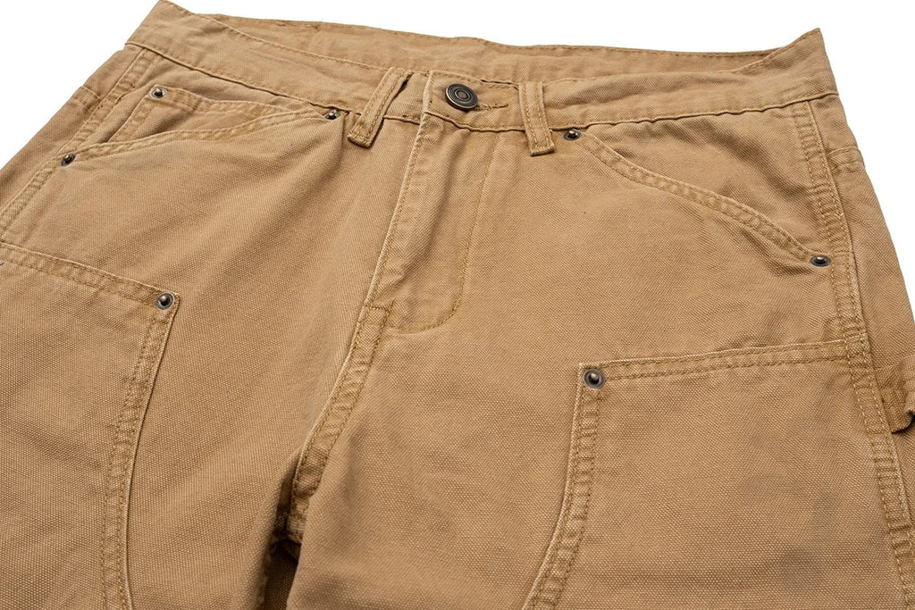 Close-up of tan canvas work pants showing the waistband, belt loops, button, and front pockets on a white background.