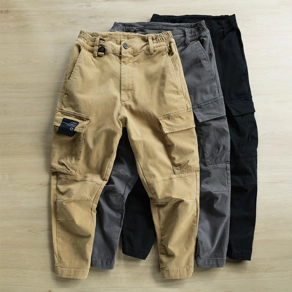 Three pairs of cargo pants laid flat on a light wood surface, with the front pair in tan and the pairs behind it in dark gray and black.