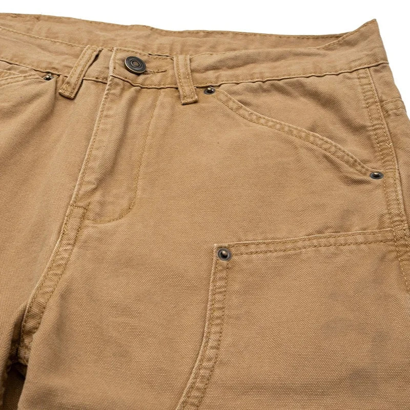 Close-up of tan canvas work pants showing the waistband, belt loops, button, front pockets, and rivets.