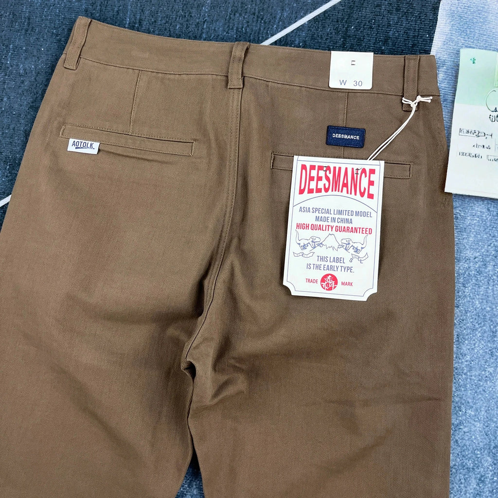 A pair of brown chino pants shown from the back, featuring a paper tag on the pocket that reads 'DEESMANCE' and a small 'FUGU' label on the side pocket, laid flat on a blue textured surface.