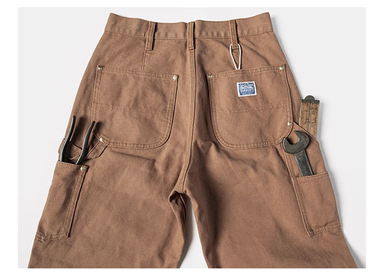 Brown canvas work pants shown from the back, featuring utility pockets with tools like a wrench and pliers, and a small blue brand patch on the back pocket.