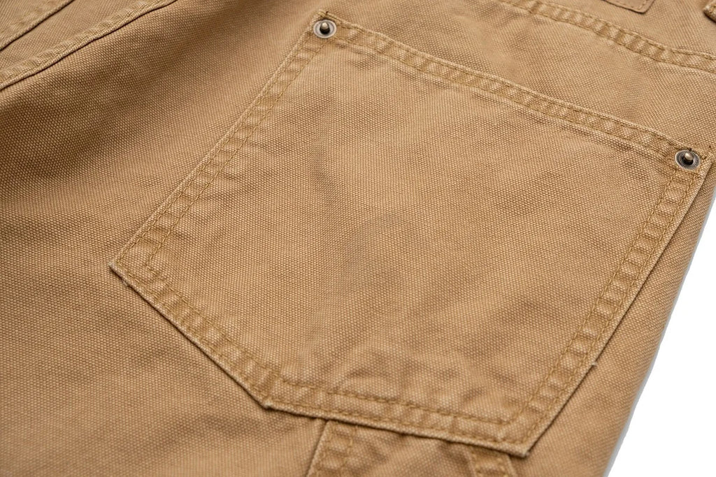 Close-up of a tan canvas garment showing a front patch pocket with metal rivets and stitched seams.