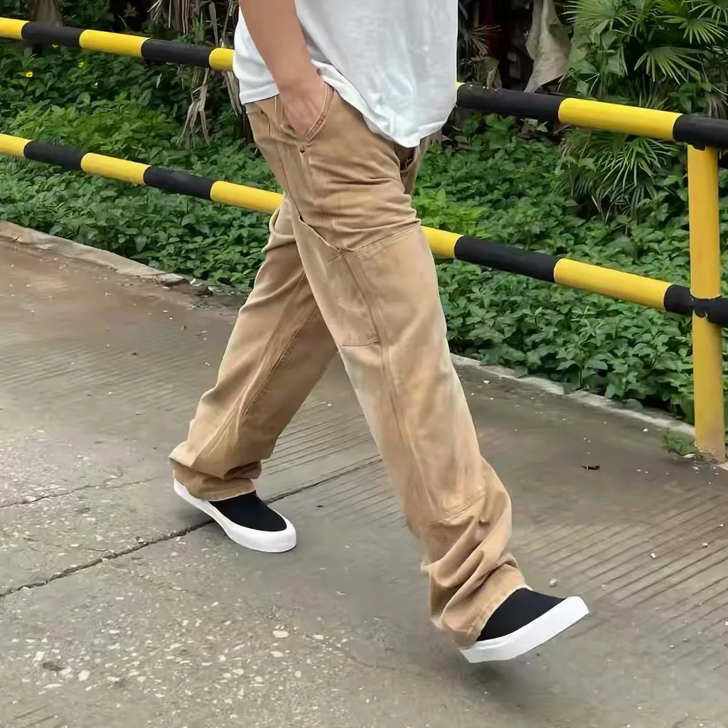 A person walking outdoors wearing loose beige corduroy pants, a white t-shirt, and black slip-on shoes with white soles, with a yellow and black railing and green foliage in the background.