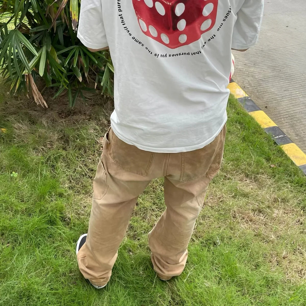 A person standing outdoors on grass, shown from the back wearing a white graphic T-shirt with a large red die print and tan cargo pants.
