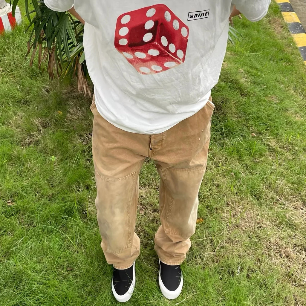 A person standing on grass wearing a white t-shirt with a large red die graphic and the word 'saint' in a black box, paired with tan cargo-style pants and black slip-on shoes.