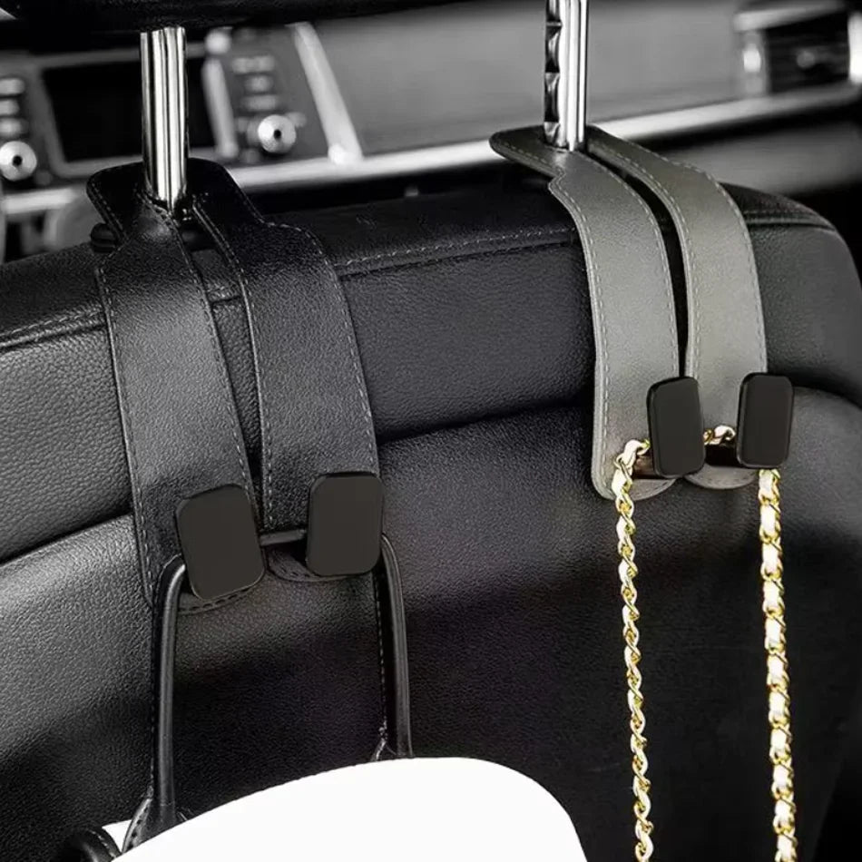 A pair of black car headrest organizer clips attached to the back of a black leather car seat, holding two car keys and a gold chain bag strap.