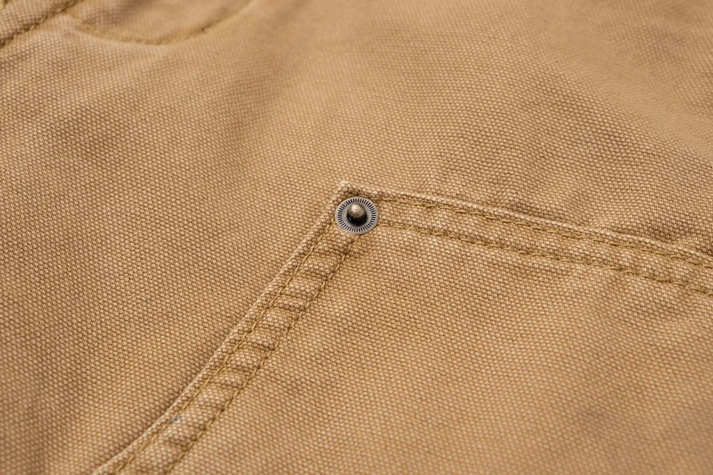 Close-up of a tan canvas garment showing a diagonal seam, a pocket, and a small metal snap button.