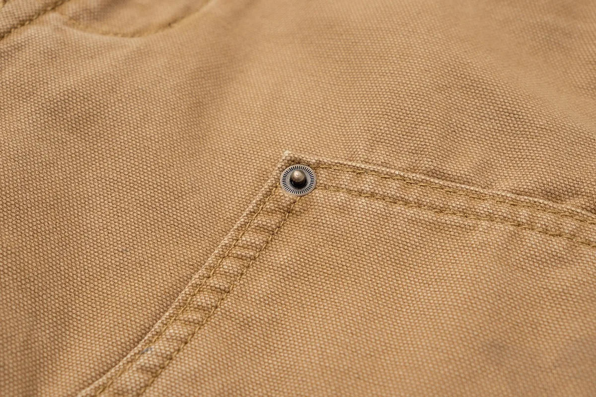 Close-up of a tan canvas garment showing a diagonal seam, a pocket, and a small metal snap button.