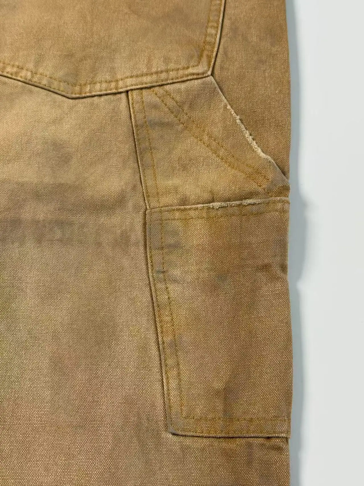 Close-up of a tan canvas or denim-like fabric garment showing a pocket seam and stitched texture against a light gray background.