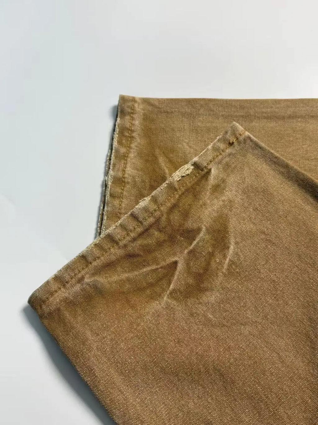 A close-up of folded tan or camel-colored fabric, showing a textured woven material and stitched edges, laid on a plain light background.