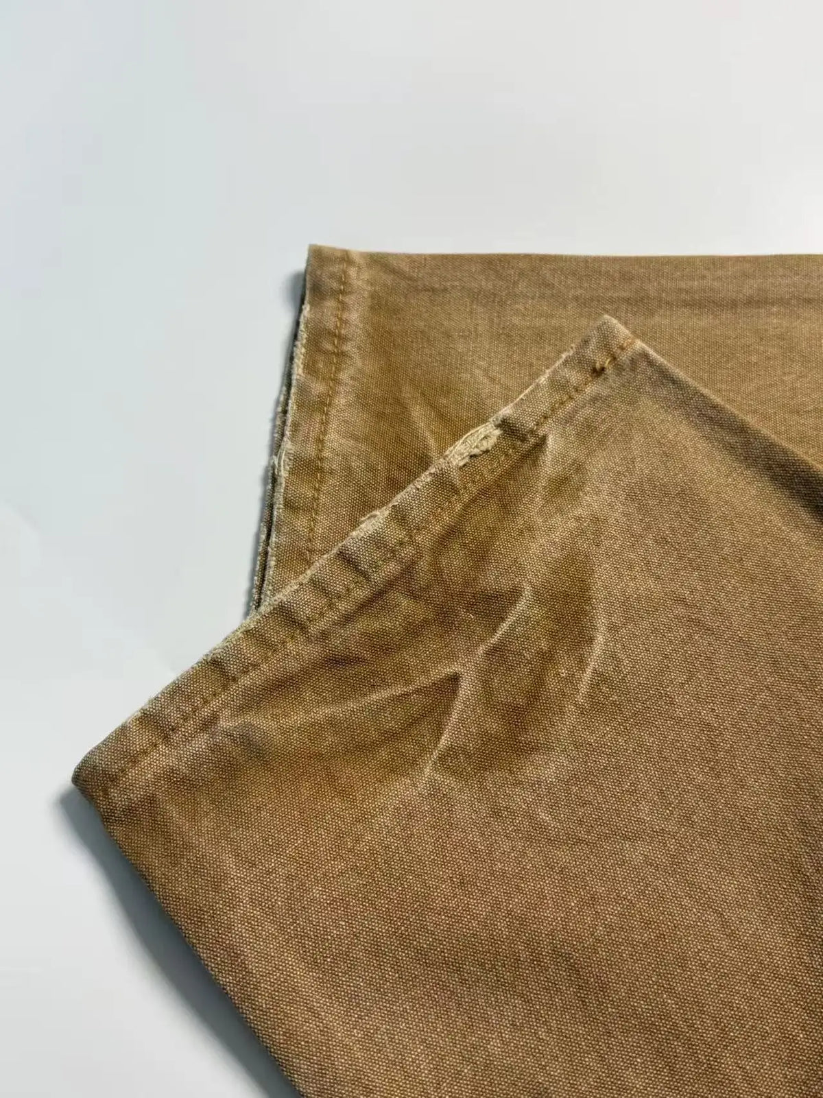 A close-up of folded tan or camel-colored fabric, showing a textured woven material and stitched edges, laid on a plain light background.