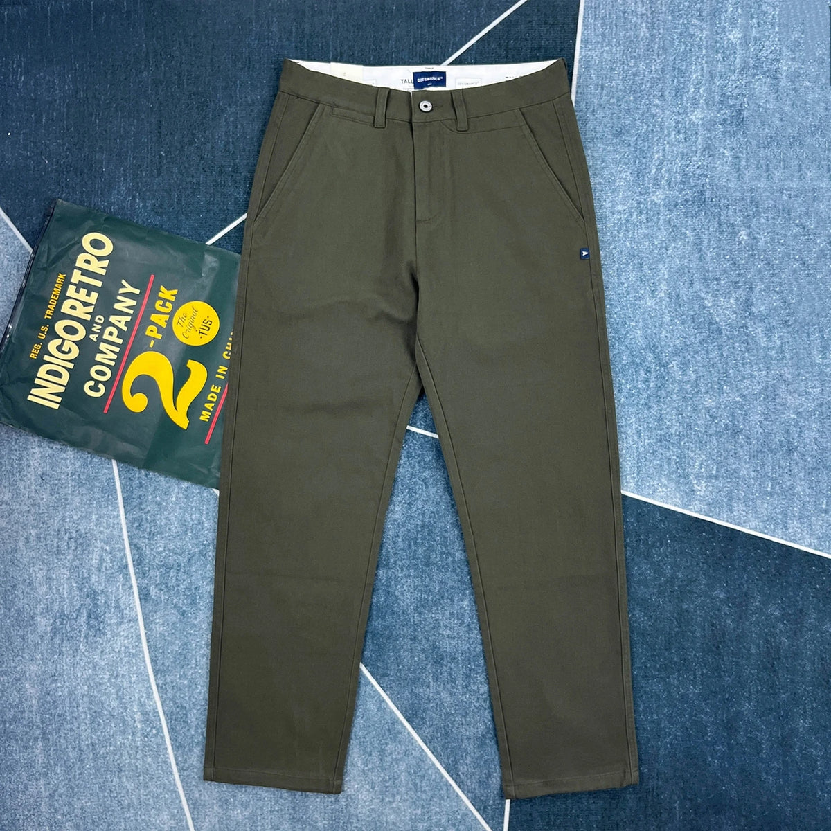 A pair of olive green chino pants laid flat on a blue patterned background, with a branded green hanger tag placed beside them. The pants have a button closure, belt loops, side pockets, and a small blue brand tab near the pocket.