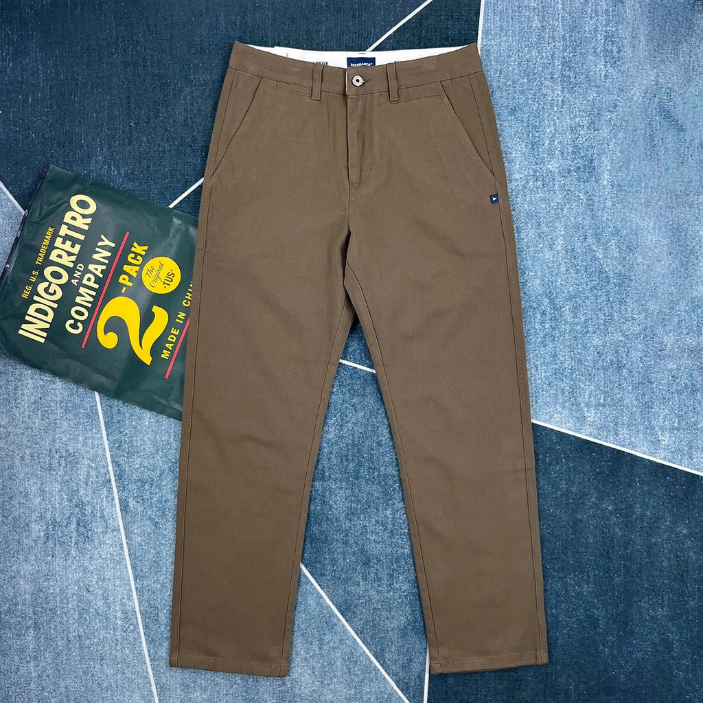 A pair of brown chino pants laid flat on a blue textured background next to a green 'Indigo Retro Company 2-Pack' label, showing the brand tag at the waistband.