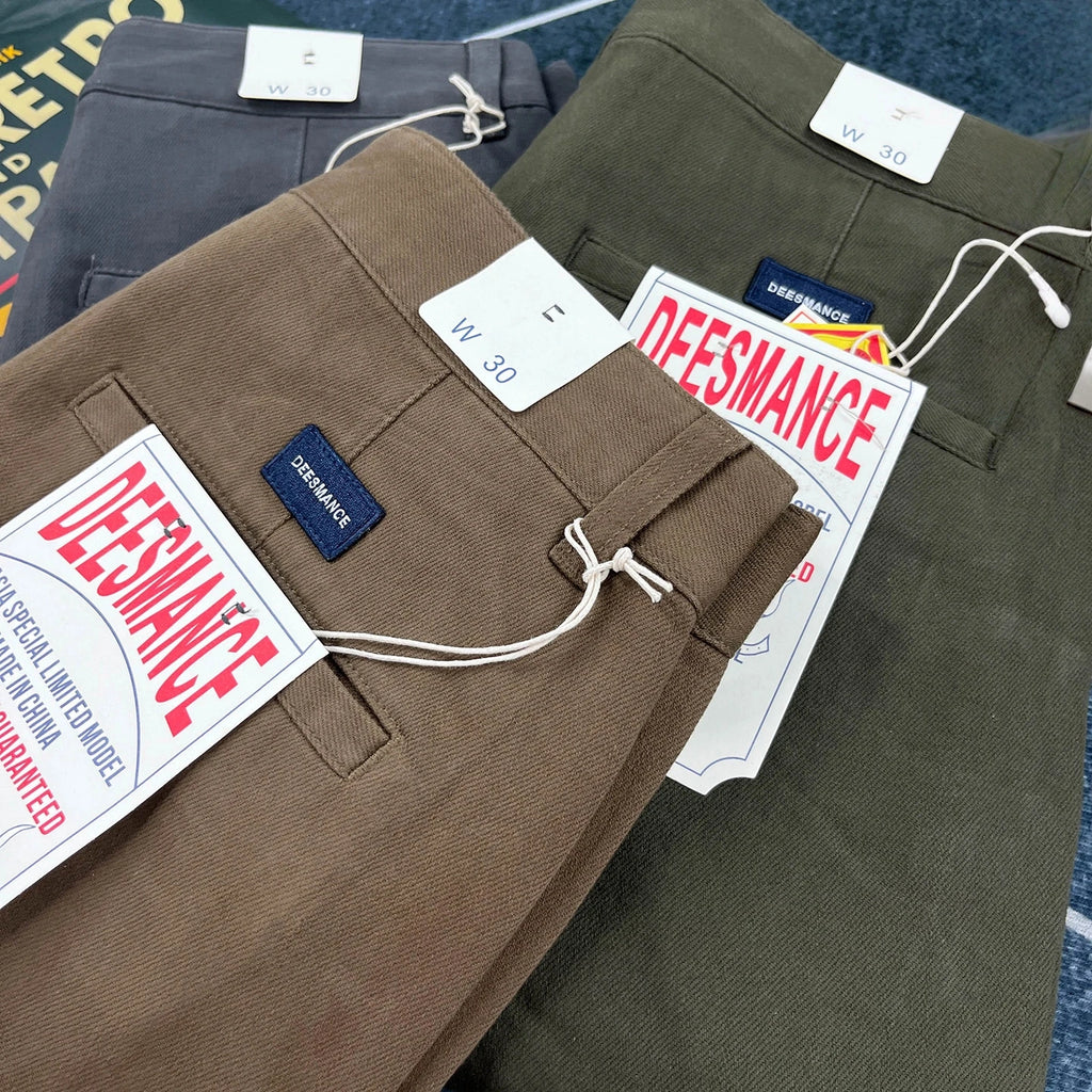 Three pairs of folded pants stacked on top of each other: a brown pair in front, an olive green pair behind it, and a dark gray pair at the top. Each pair has white size tags and a blue DEESMANCE brand label.