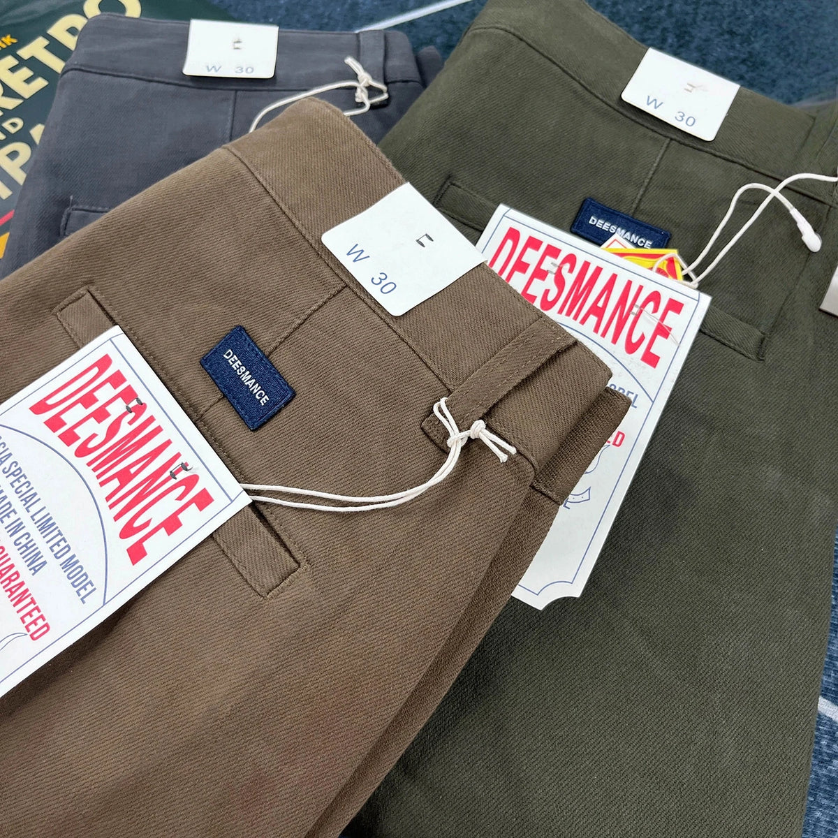 Three pairs of folded pants stacked on top of each other: a brown pair in front, an olive green pair behind it, and a dark gray pair at the top. Each pair has white size tags and a blue DEESMANCE brand label.