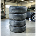 A stack of four black all-weather tires standing upright in a garage setting, with a gray car and garage equipment visible in the background.
