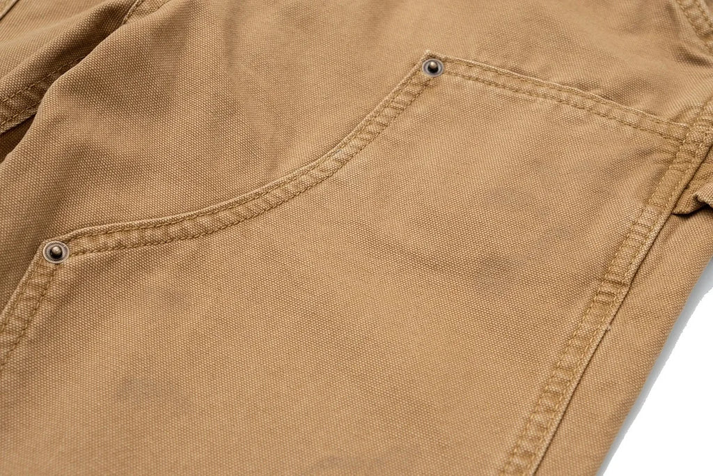 Close-up of a tan canvas garment showing a front pocket with metal rivets and stitched seams on a white background.