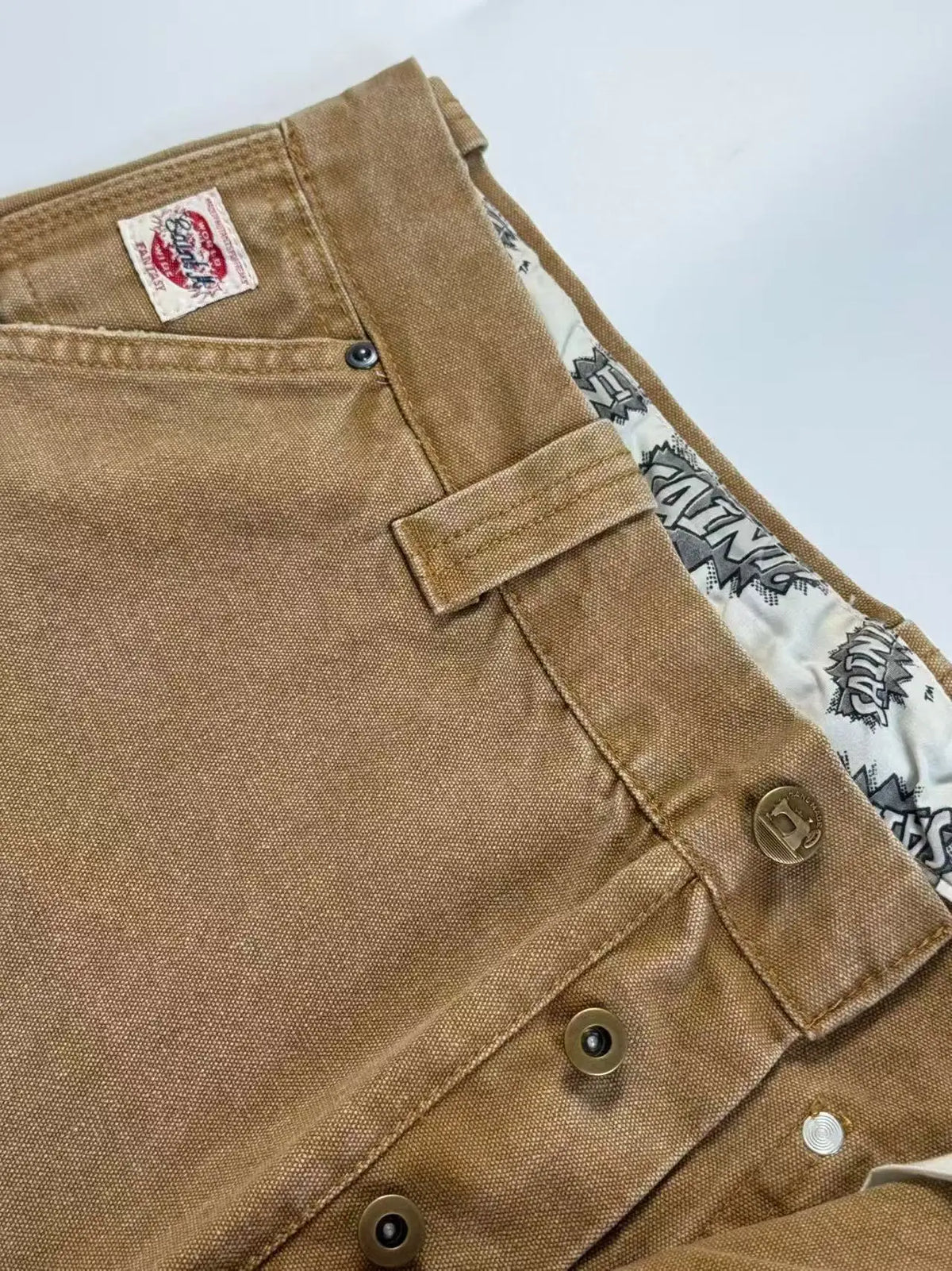 Close-up of a brown canvas utility jacket or overshirt showing the waistband, belt loops, button closure, and a patterned inner lining.