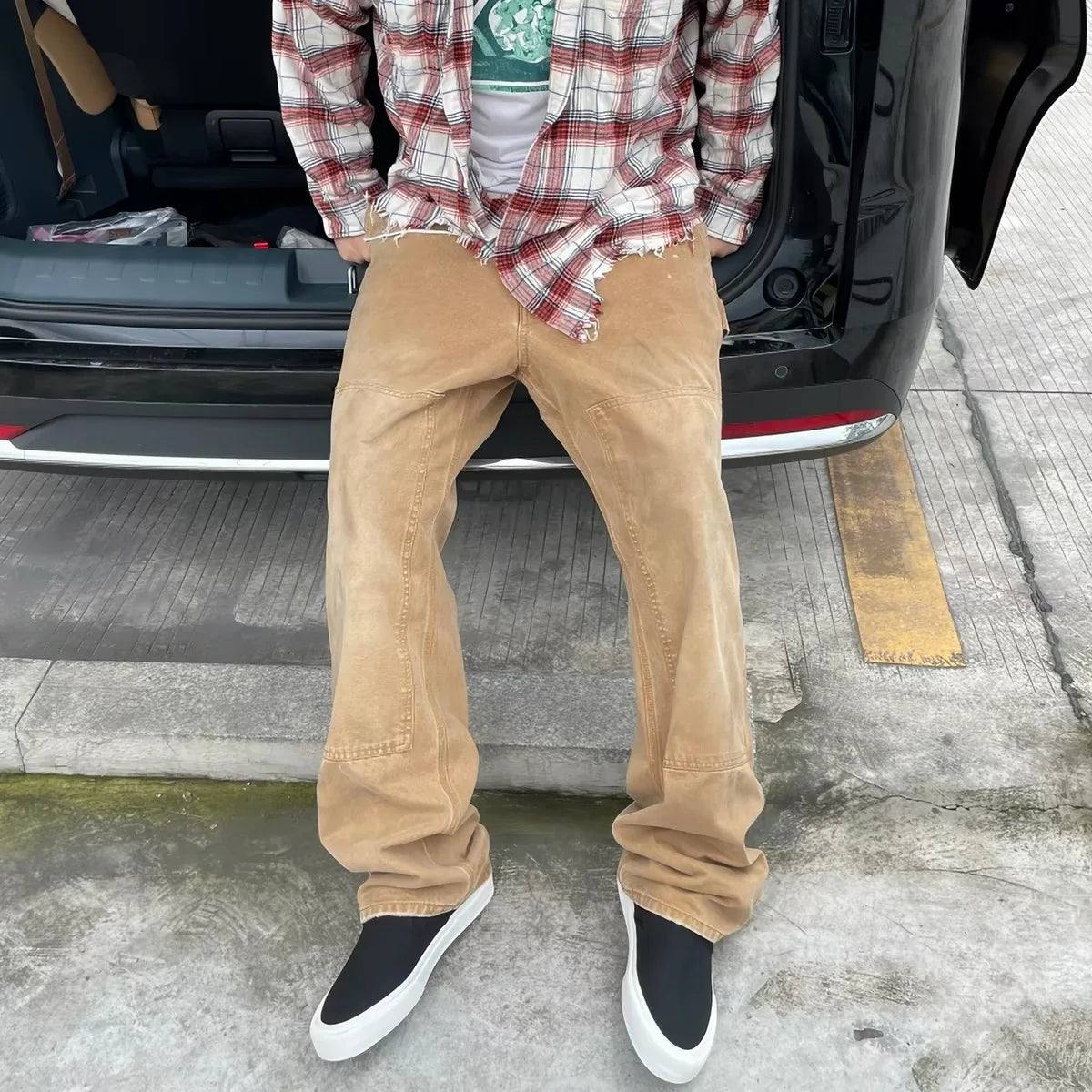 A person sitting on the open trunk of a black SUV, wearing tan corduroy pants, a red and white plaid shirt, and black slip-on shoes with white soles.