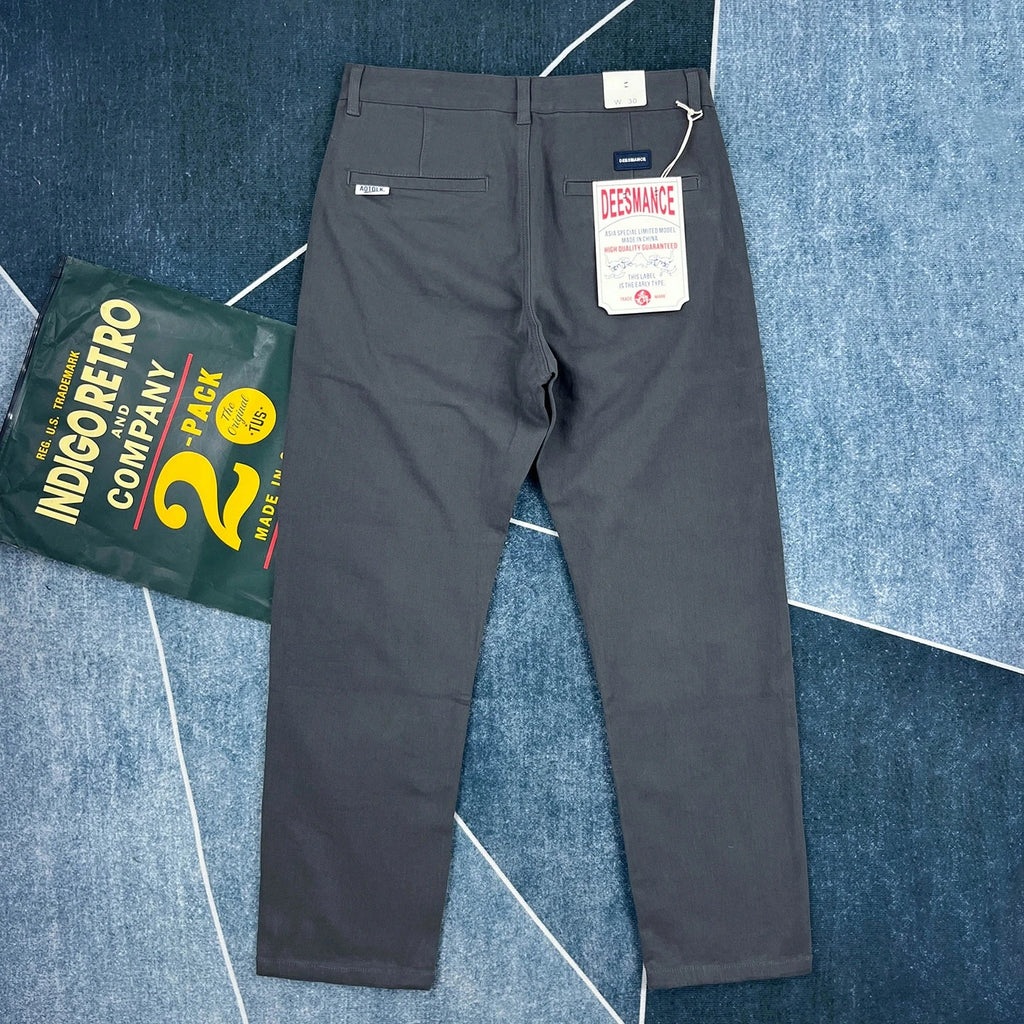 A pair of dark gray chino pants laid flat on a blue patterned rug, with a Desmanze brand tag still attached at the back waist. A green bag of IndigoFerro Company twill is partially visible on the left side.