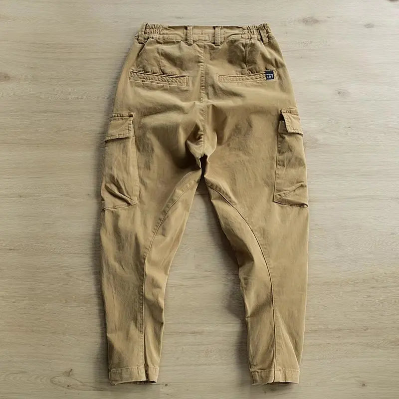 A pair of beige cargo pants laid flat on a light wood floor, showing the back pockets, side cargo pockets, and elastic waistband.