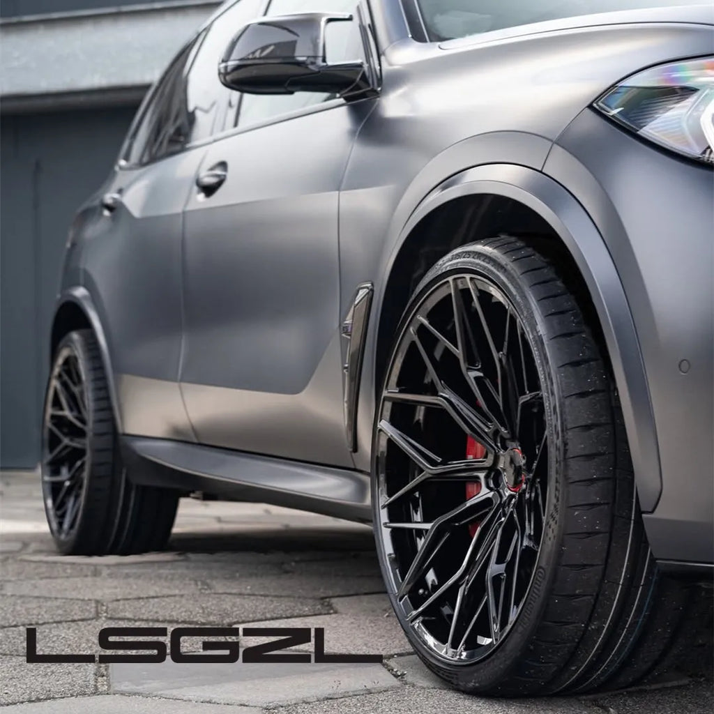 A close-up of a matte gray SUV showing the front wheel area with a large black multi-spoke aftermarket wheel and tire. The vehicle is photographed outdoors on a paved surface with a building wall in the background.