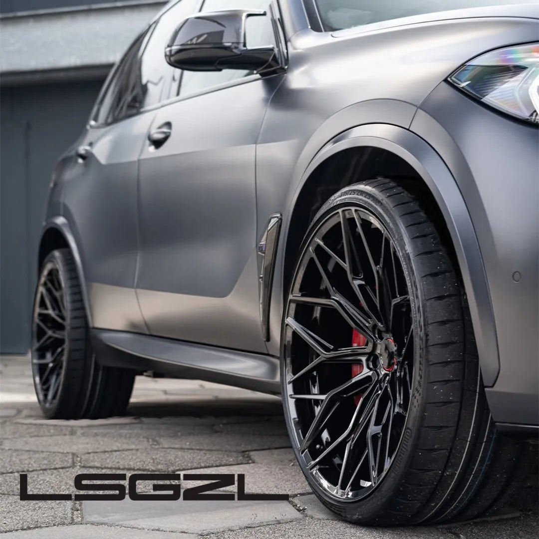 A close-up of a matte gray SUV showing the front wheel area with a large black multi-spoke aftermarket wheel and tire. The vehicle is photographed outdoors on a paved surface with a building wall in the background.