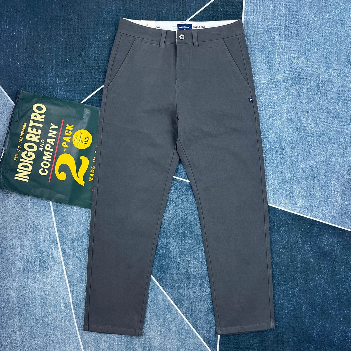 A pair of dark gray slim-fit chino pants laid flat on a blue patterned background, with a green Indigoretro Company bag placed to the left.
