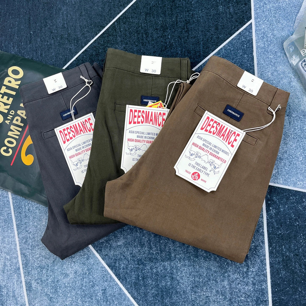 Three pairs of folded Deesmance trousers in dark gray, olive green, and brown, arranged diagonally on a blue patterned surface with product tags visible.