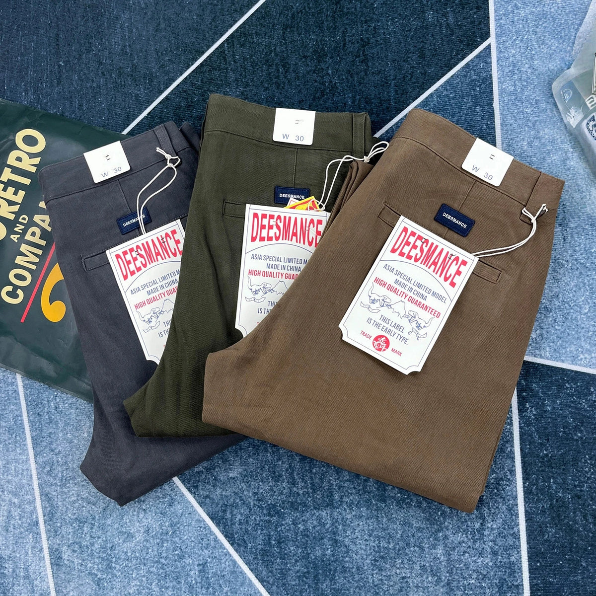 Three pairs of folded Deesmance trousers in dark gray, olive green, and brown, arranged diagonally on a blue patterned surface with product tags visible.