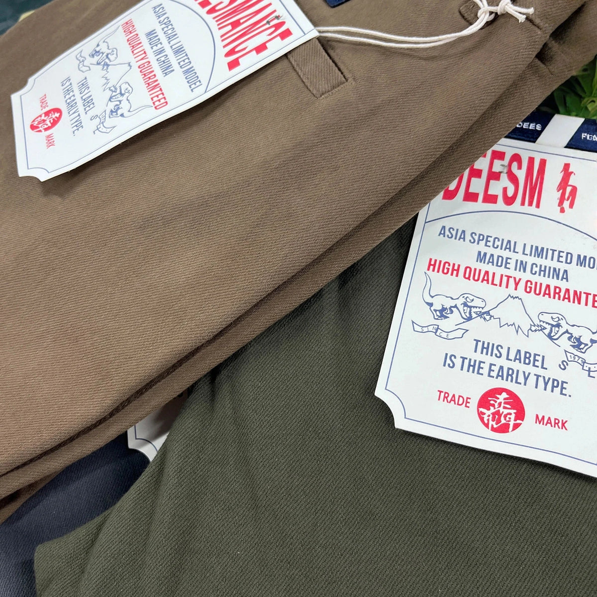 Close-up of folded clothing showing two garments, one in taupe-brown and one in olive green, both with white paper tags printed with red and blue text and graphics.