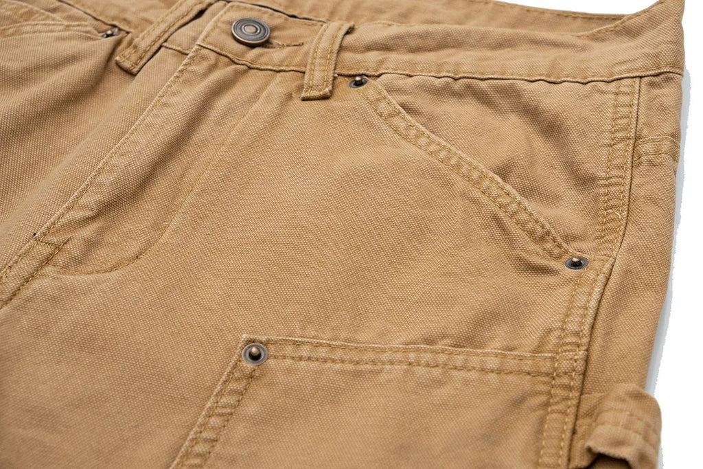 Close-up of tan canvas work pants showing the waistband, belt loops, front pocket, and rivets on a white background.