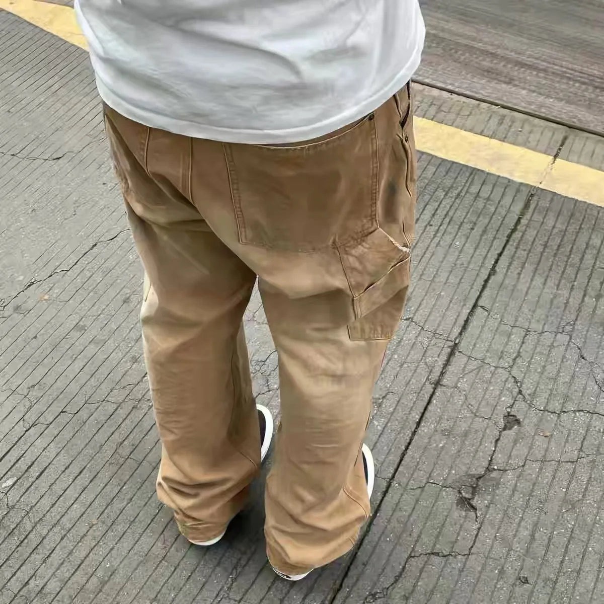 A person viewed from behind wearing tan cargo pants, a white t-shirt, and white sneakers, standing on a gray paved street.